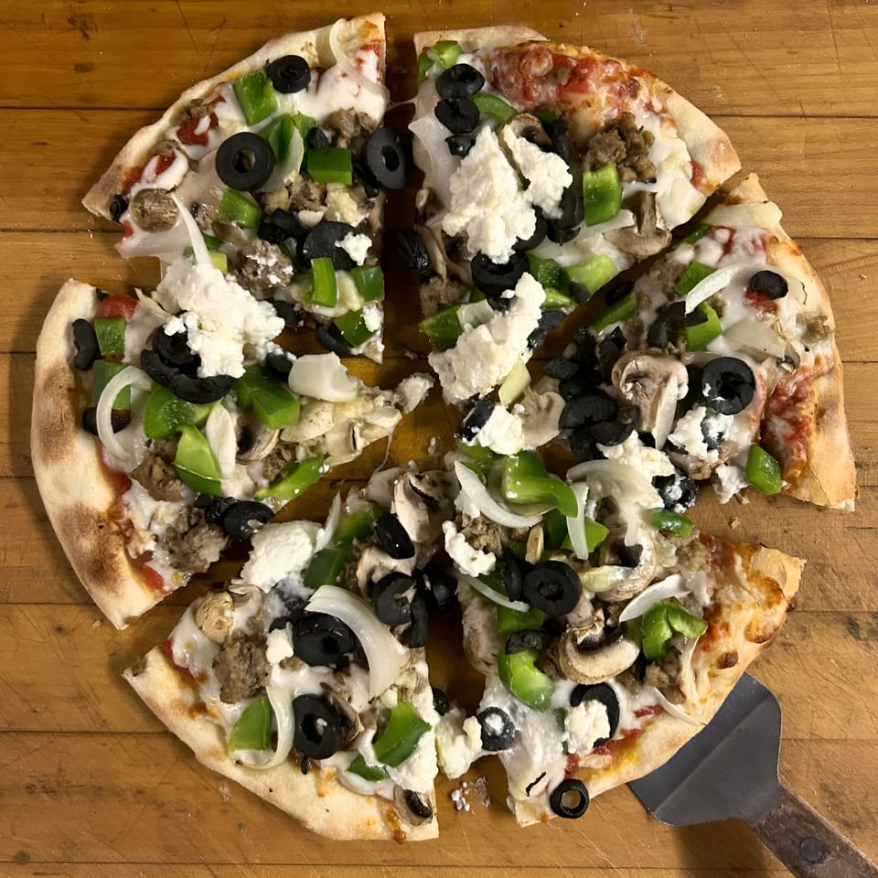 A Pacioni's Pizzeria pizza, cut into slices, with sausage, olives, peppers, onions, and ricotta.