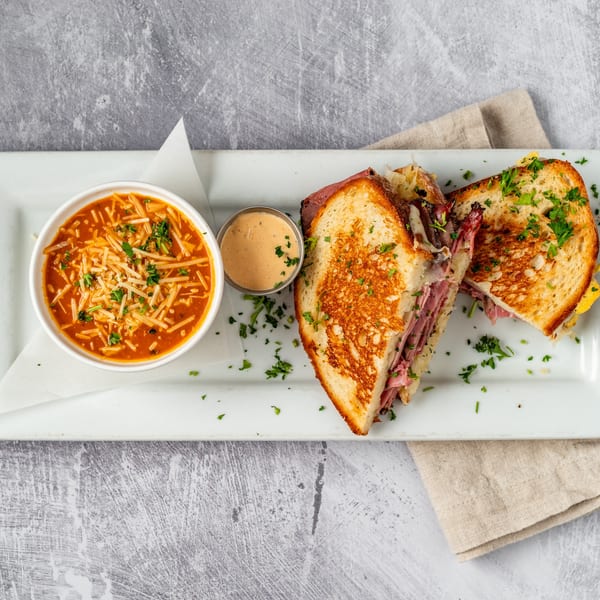 A grilled sandwich with meat and cheese, served with a bowl of tomato soup and dipping sauce.