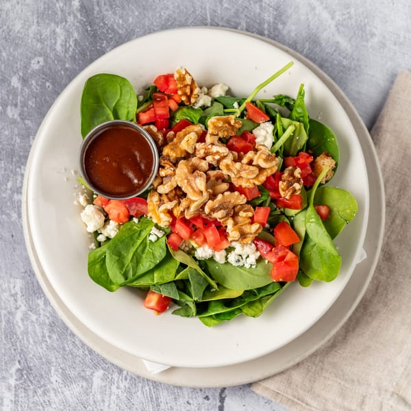 A spinach salad with walnuts, diced tomatoes, feta cheese, and a side of dressing.