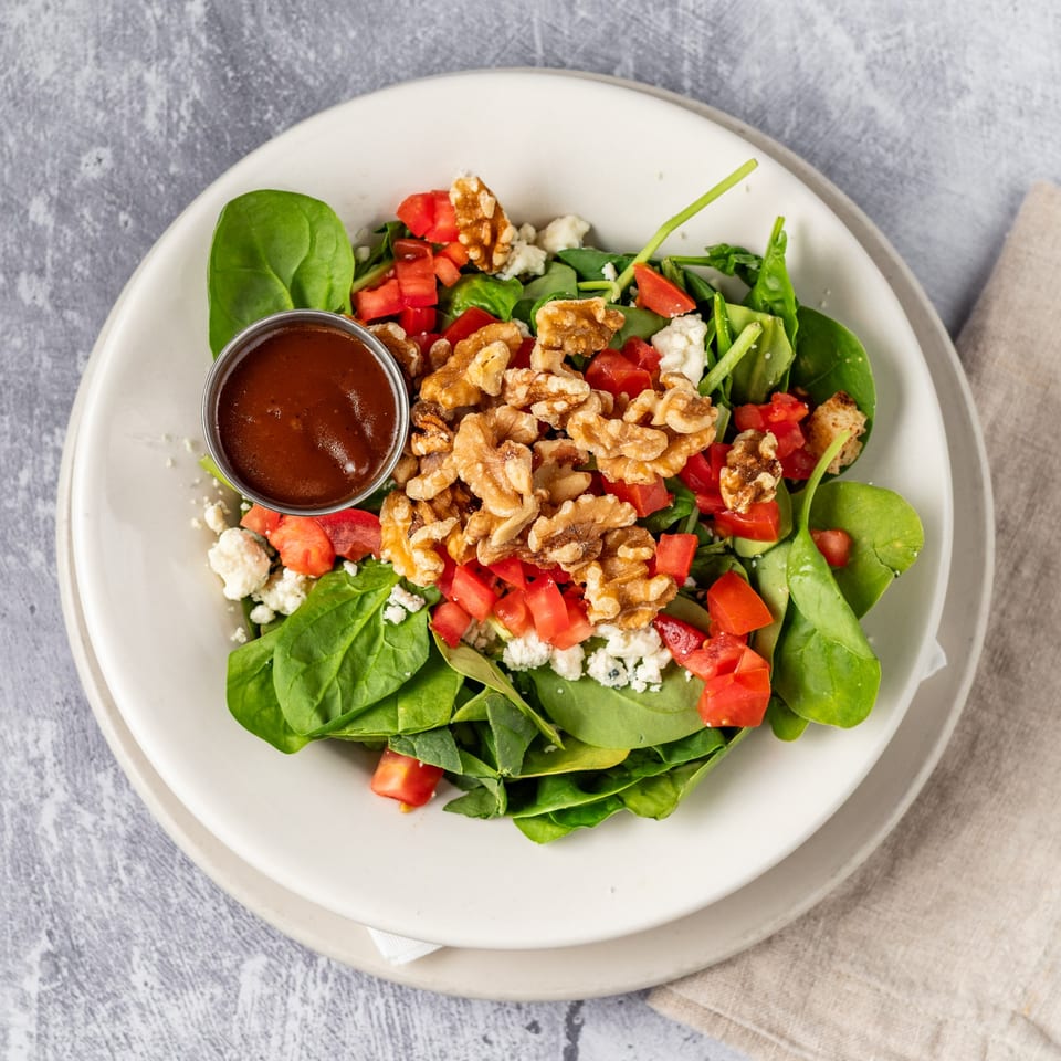A spinach salad with walnuts, diced tomatoes, feta cheese, and a side of dressing.